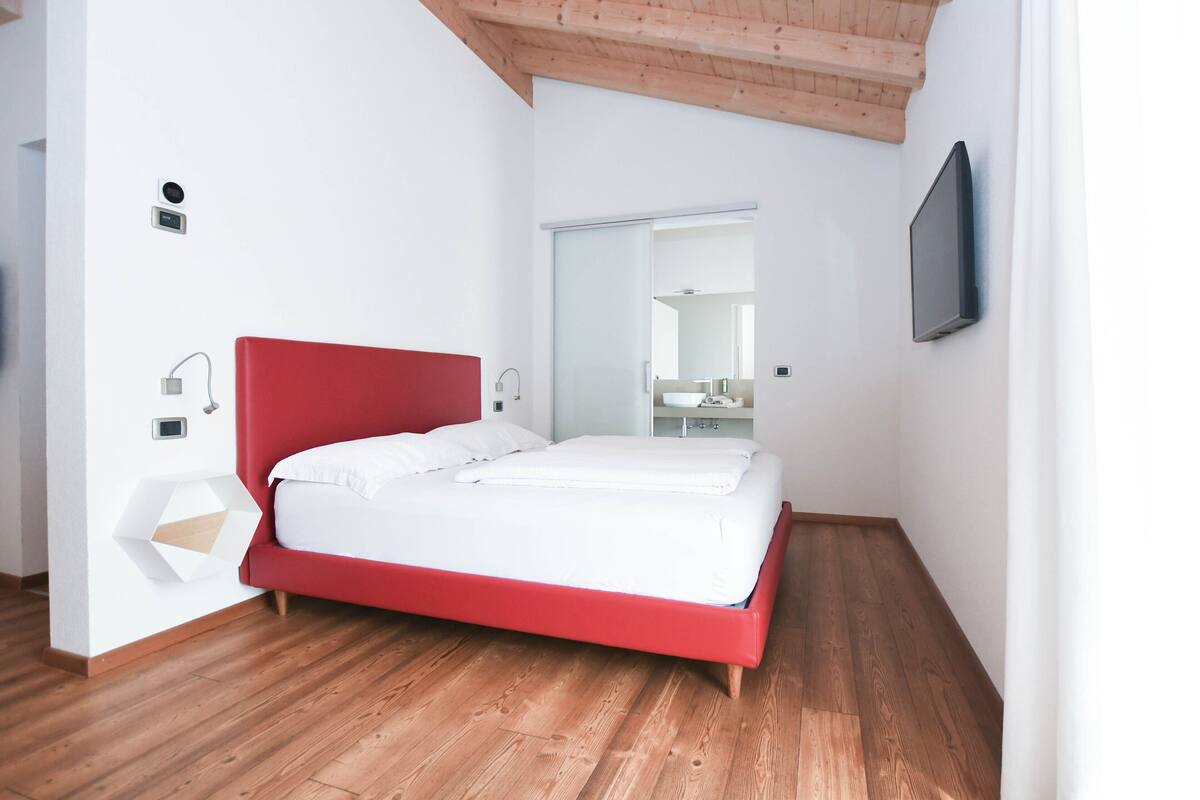 A modern bedroom with a red bed, wooden flooring, a white wall, and a flat-screen TV mounted on the wall.