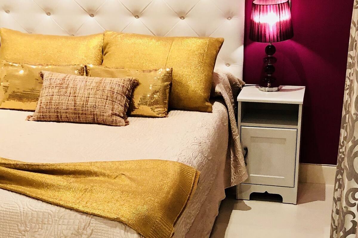 A bed with a tufted headboard, multiple pillows, a bedside table with a lamp, and a patterned curtain.
