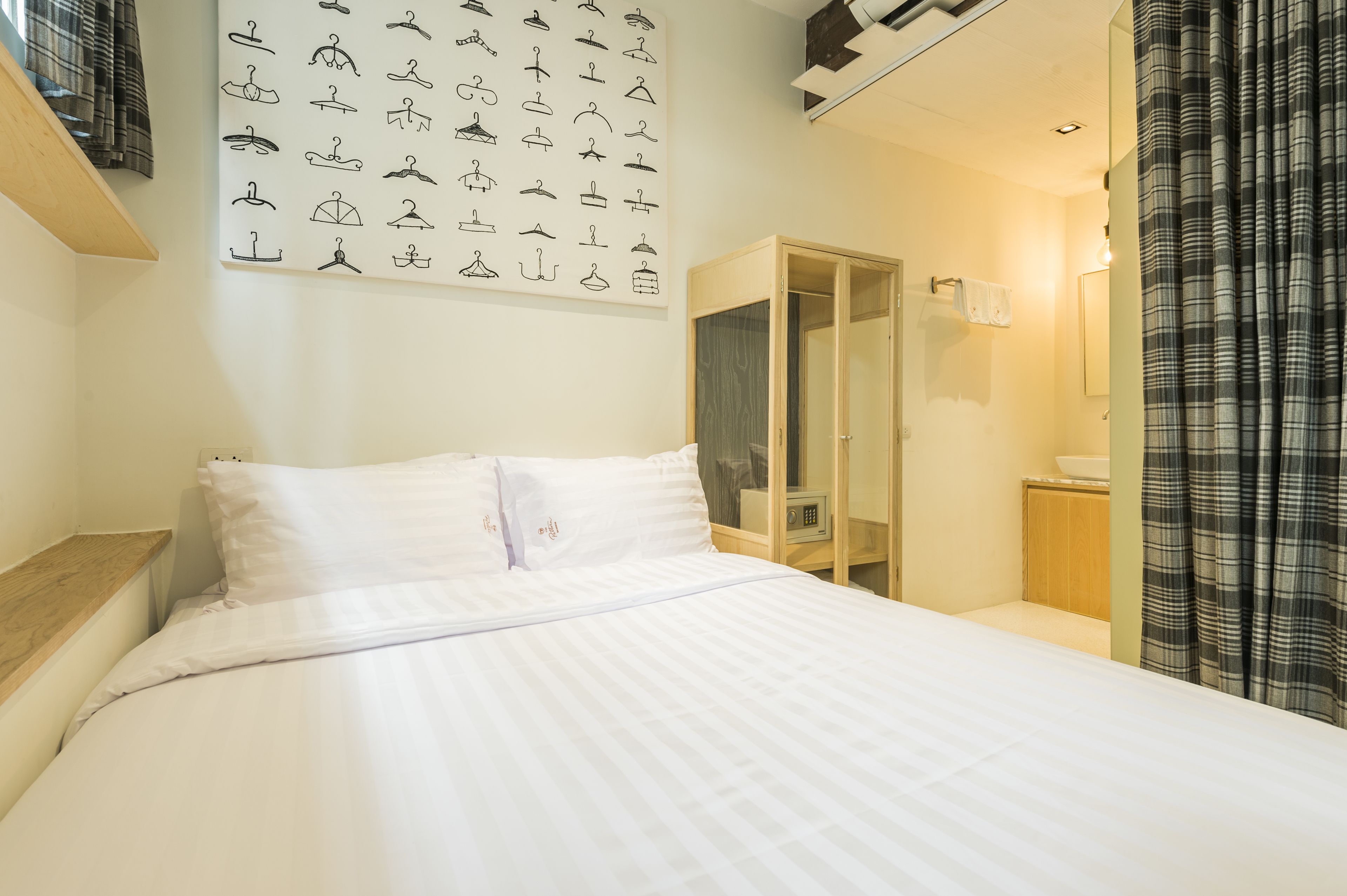 Un lit bien fait, avec du linge de lit blanc, un porte-manteau mural muni de différents cintres, une table de chevet en bois et une salle de bain avec une porte en verre et un porte-serviettes.