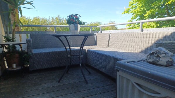 A covered outdoor seating area with a table and chairs, surrounded by potted plants and a stone feature.