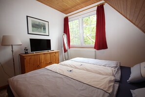 A bedroom with a bed, a TV on a wooden cabinet, a window with red curtains, and a framed picture on the wall.