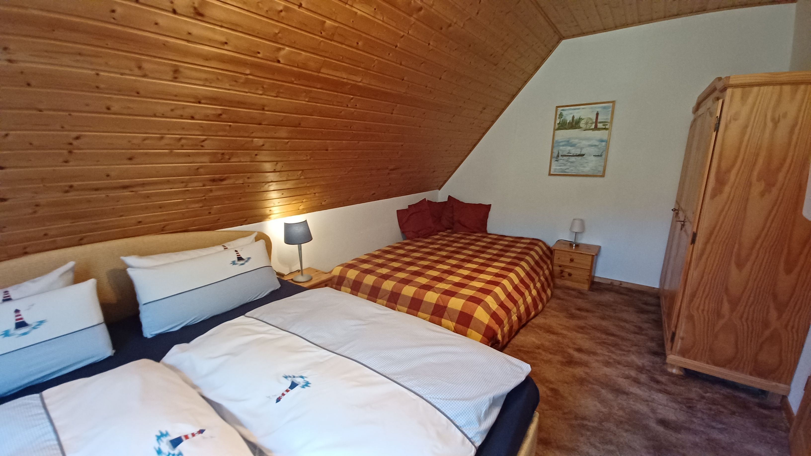 A bedroom with a sloped ceiling, wooden walls, a checkered bedspread, two bedside tables with lamps, a nightstand, and a wardrobe.