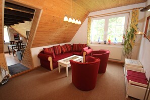 A loft-style room with a red sofa, white coffee table, and red chairs. There is a window with curtains, a plant, and a radiator.