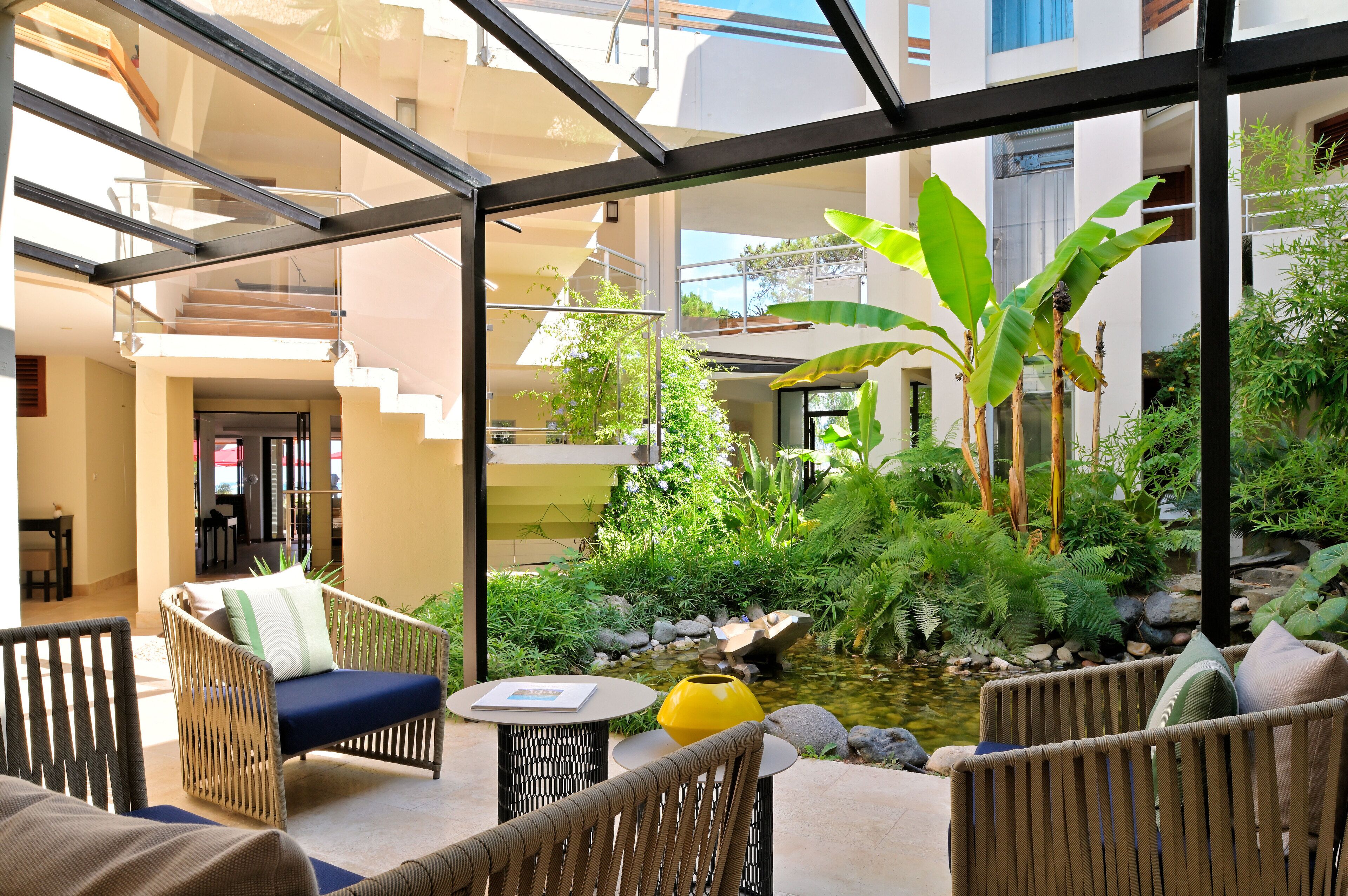 Un jardin intérieur moderne avec un petit étang, des meubles en osier et un escalier clos par des parois de verre.
