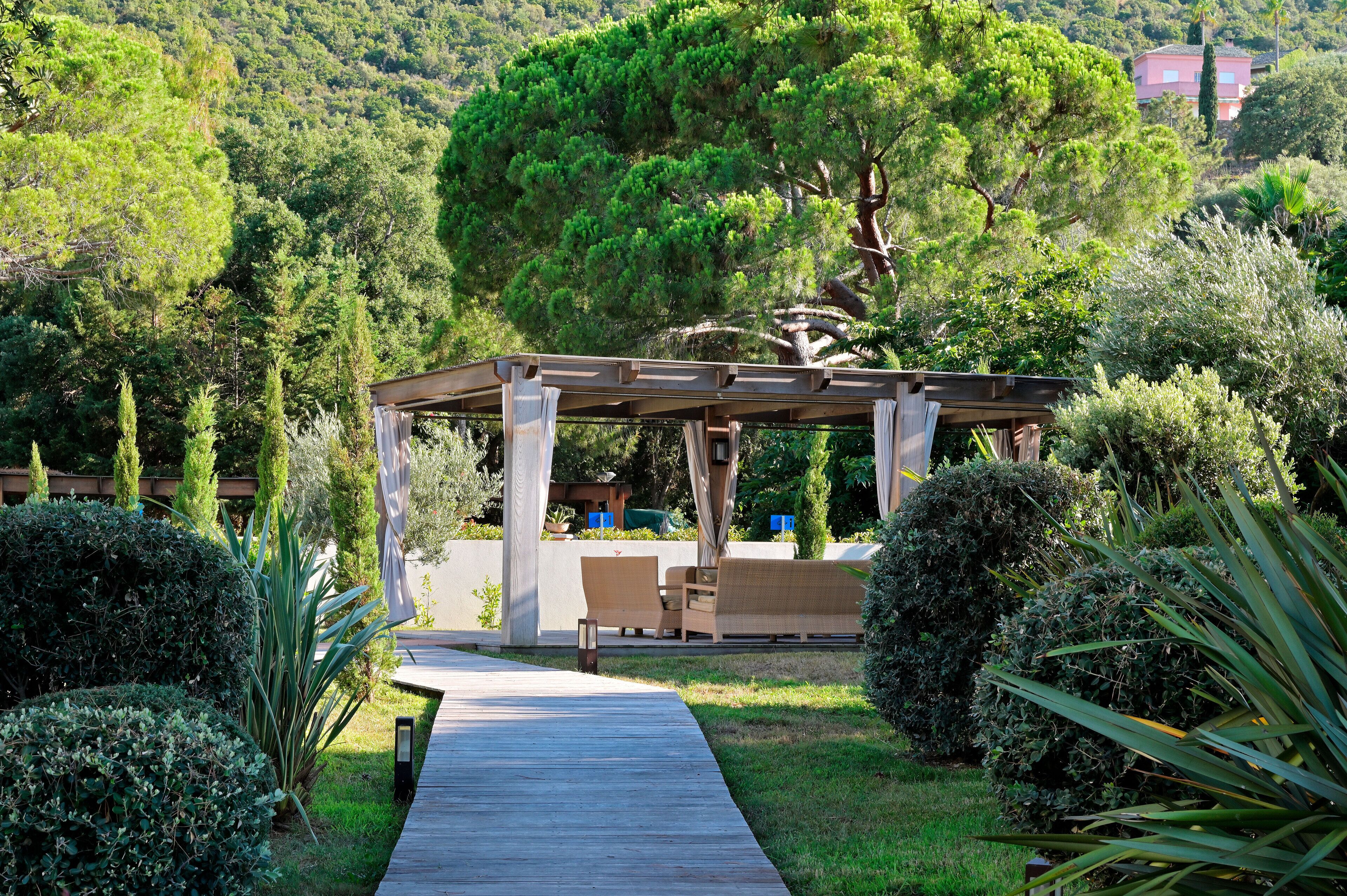 A wooden deck leading to a covered seating area with a pergola, surrounded by lush greenery and a mountainous backdrop.