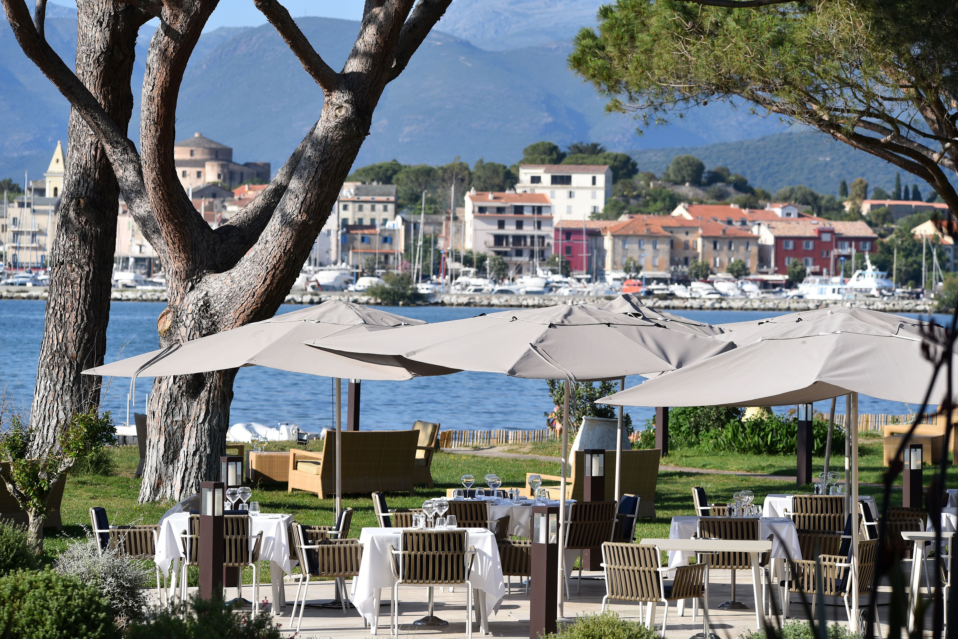 An outdoor dining area with tables and umbrellas, overlooking a marina with boats and buildings in the background.