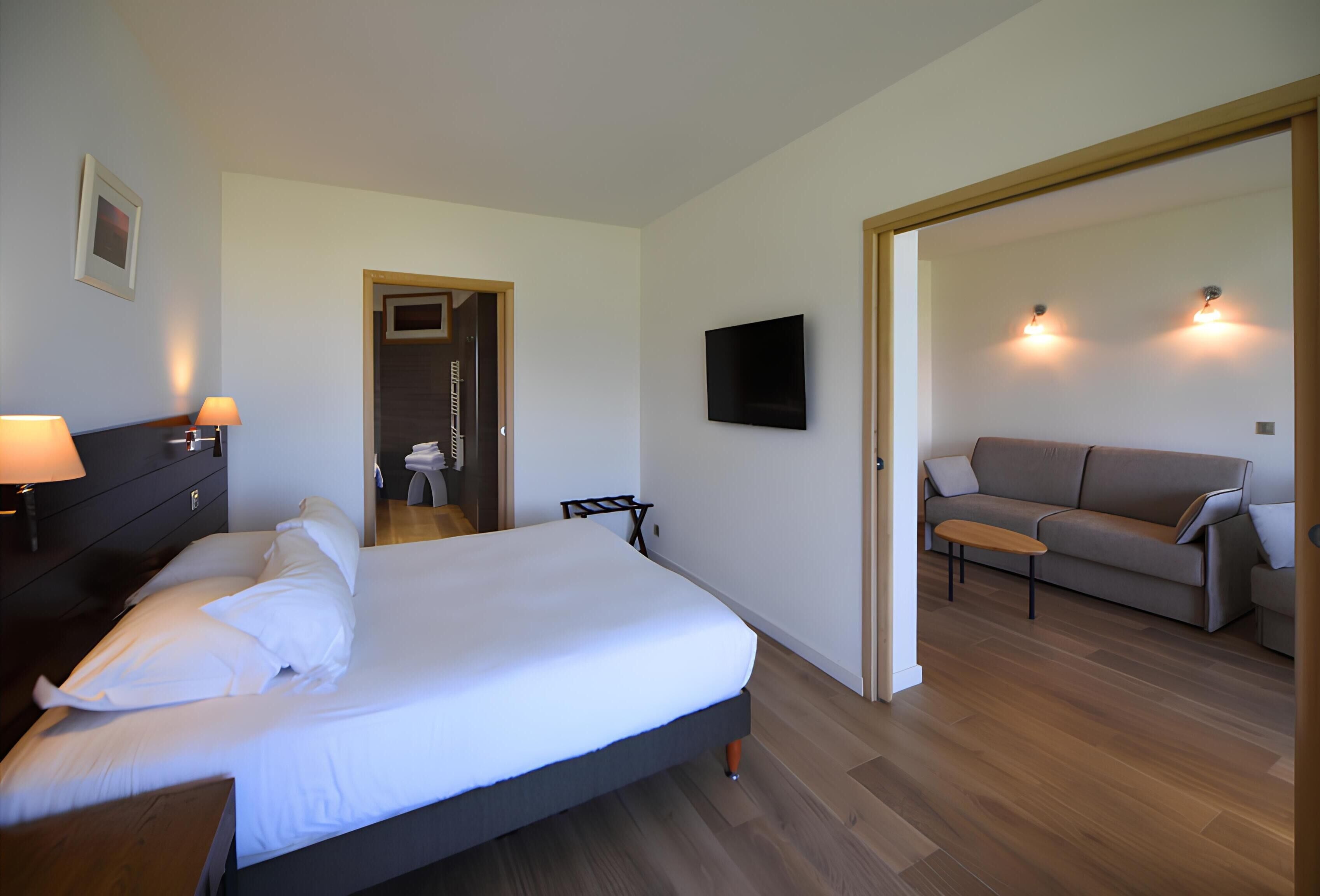 A hotel room with a bed, bedside table, lamp, TV, and a sofa in the adjacent living area.