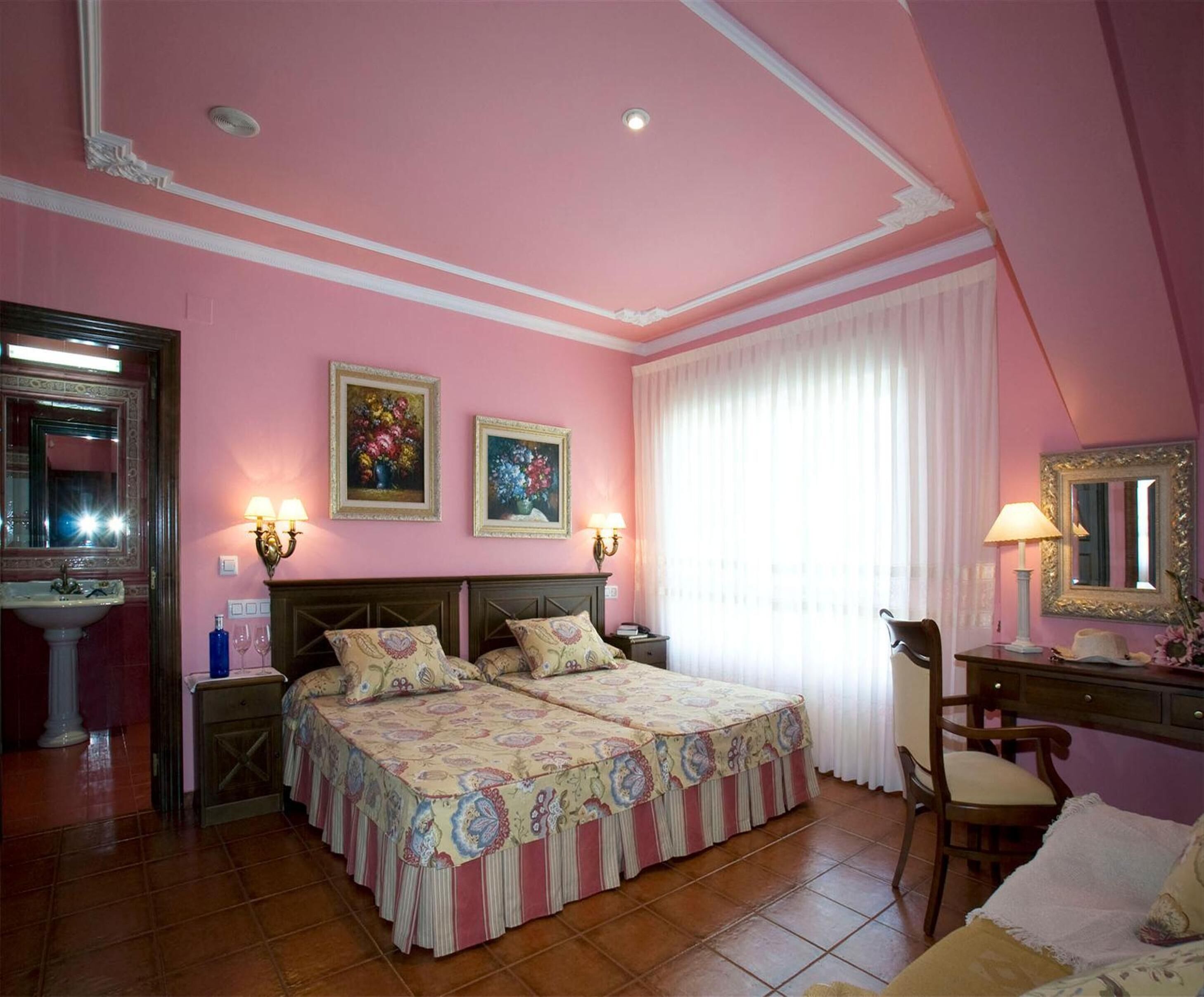 Une chambre décorée dans des tons de rose, avec deux lits, une coiffeuse avec un miroir et une fenêtre ornée de rideaux légers.