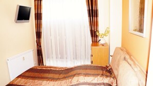 A bedroom with a bed, a bedside table with a lamp, a window with curtains, and a wall-mounted TV.