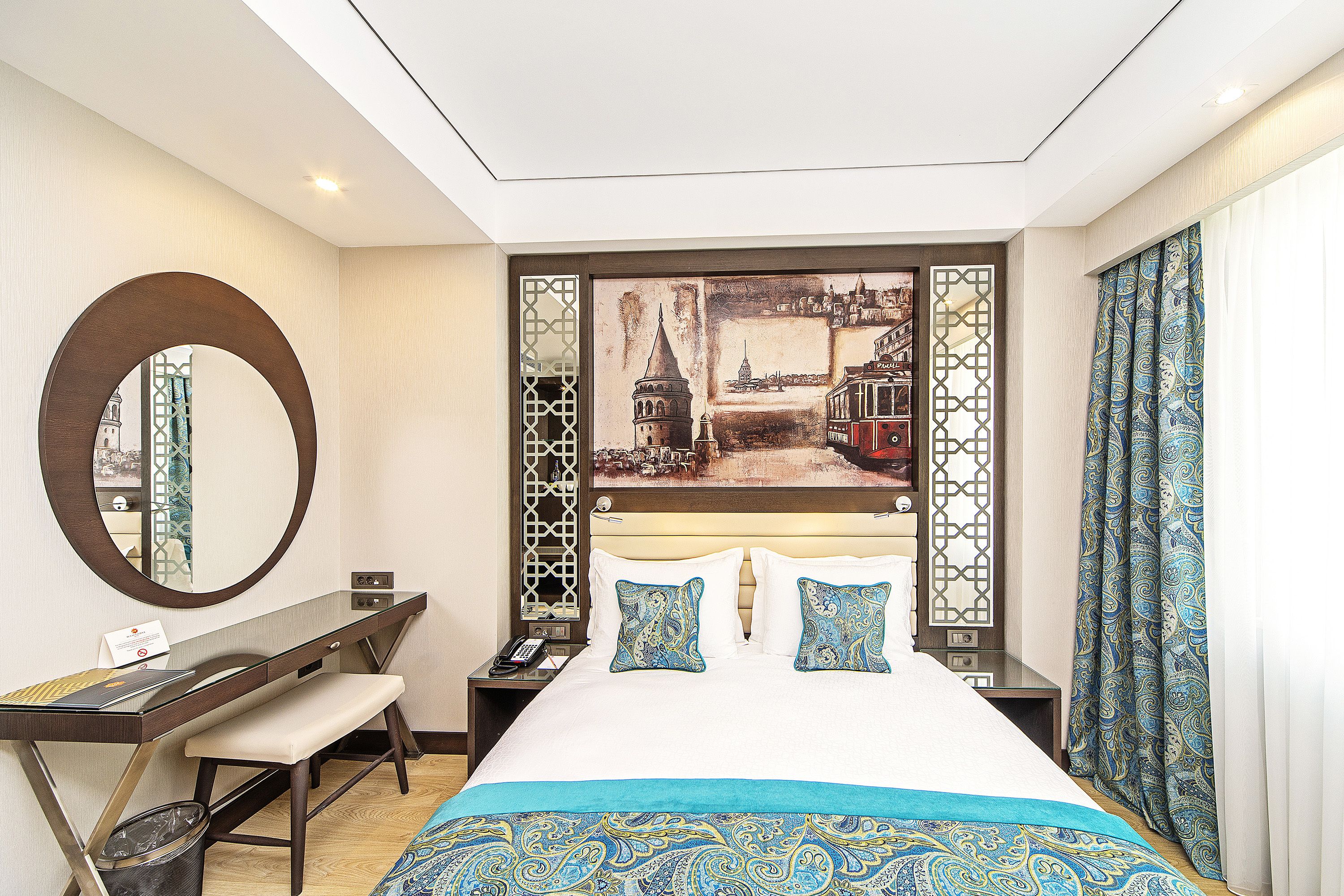 Habitación de hotel con cama, escritorio, silla, espejo, un cuadro en la pared y ventana con cortinas.