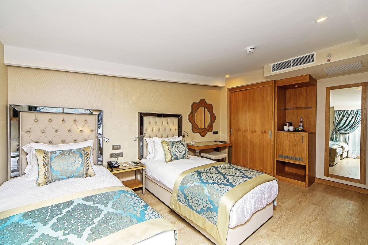 A hotel room with two beds, a wooden wardrobe, a mirror, and a small table with a lamp.