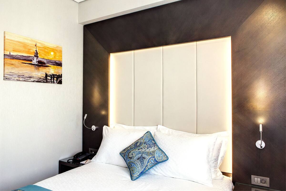A hotel room with a bed, a nightstand, a wall-mounted phone, and a painting of a sunset over a lighthouse.