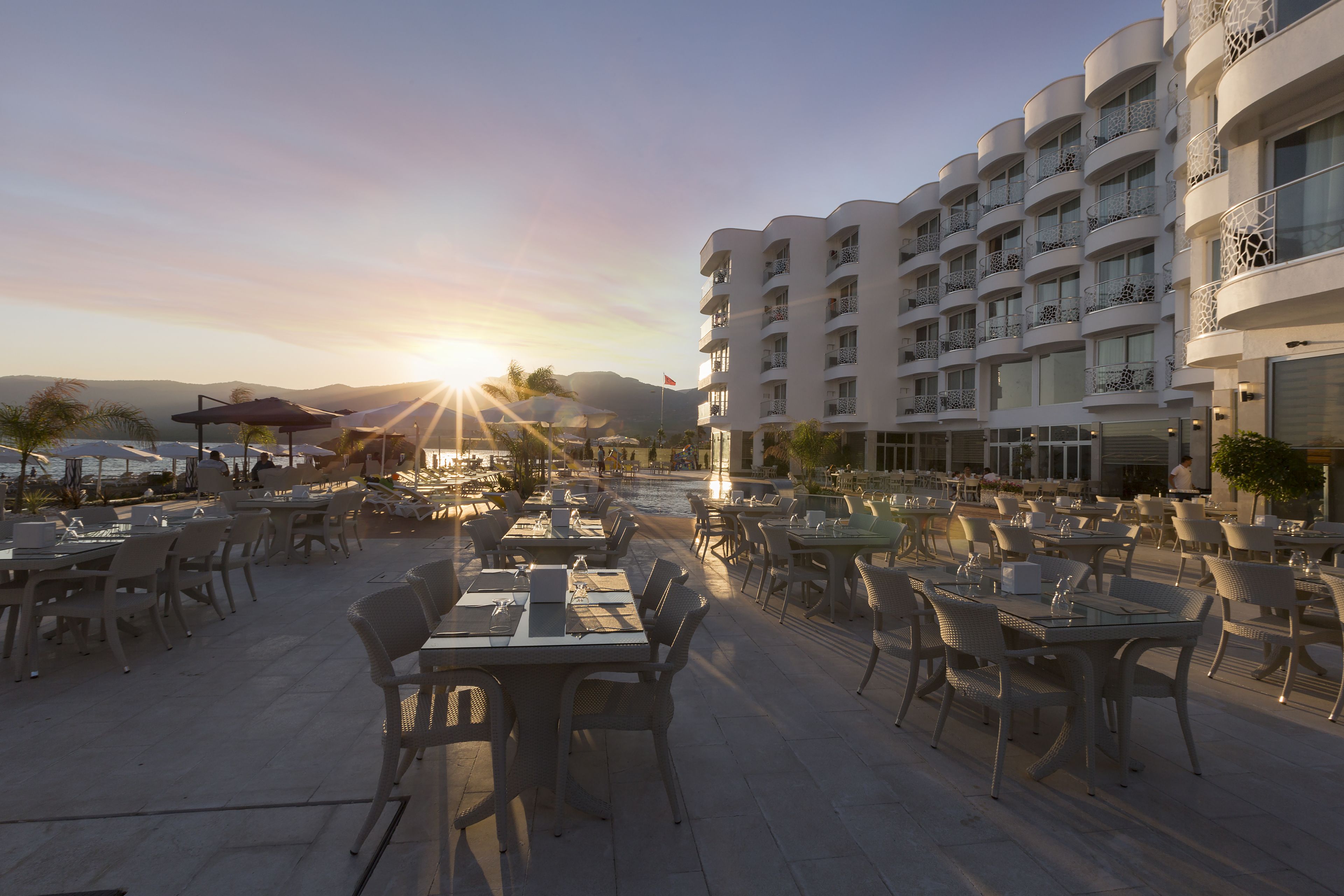 An outdoor dining area with tables and chairs, a building with balconies, and a sunset in the background.