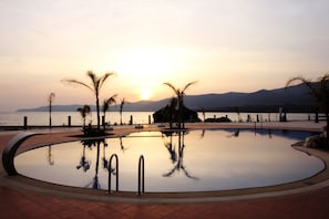 Une piscine avec vue sur le coucher de soleil, des palmiers et une étendue d’eau en arrière-plan.
