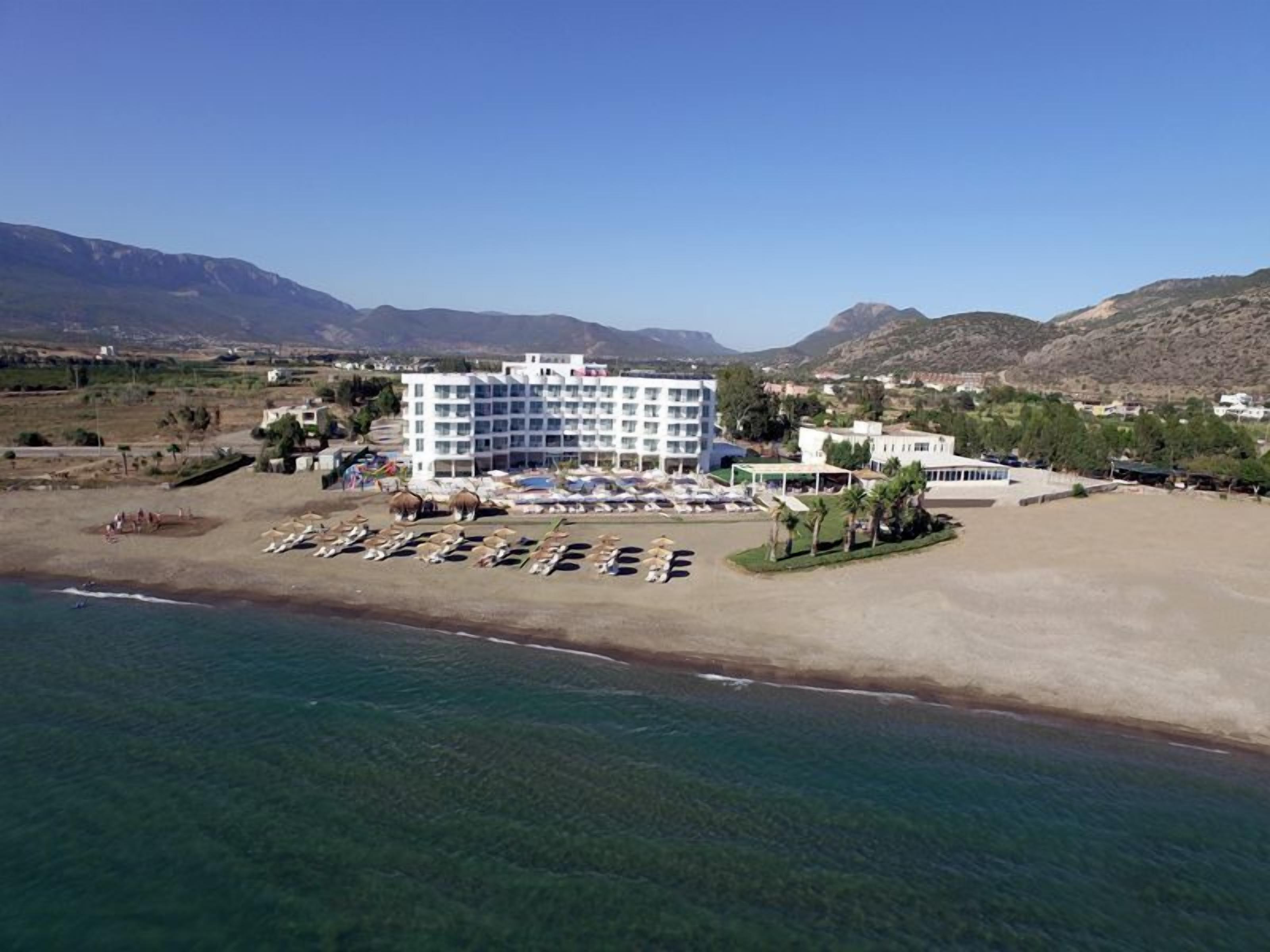 Een hotel aan de kust met meerdere verdiepingen, een strandgedeelte met parasols, en een parkeerplaats.