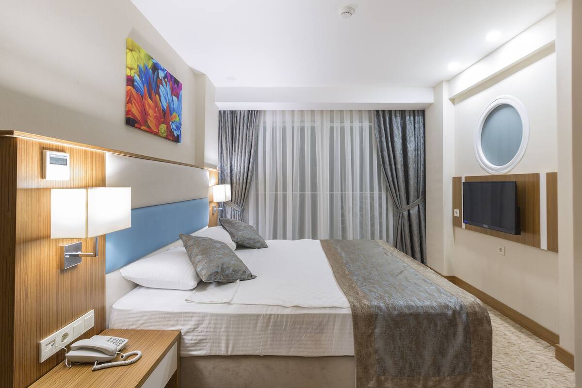 Habitación de hotel con cama, mesita de noche con teléfono, lámpara, un cuadro, televisor y cortinas.