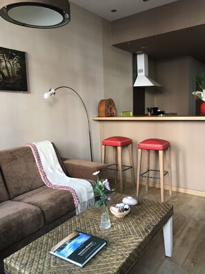 Un salon avec un canapé marron, une table basse sur laquelle se trouve un livre, une lampe, et un coin cuisine avec deux tabourets rouges et une hotte blanche.