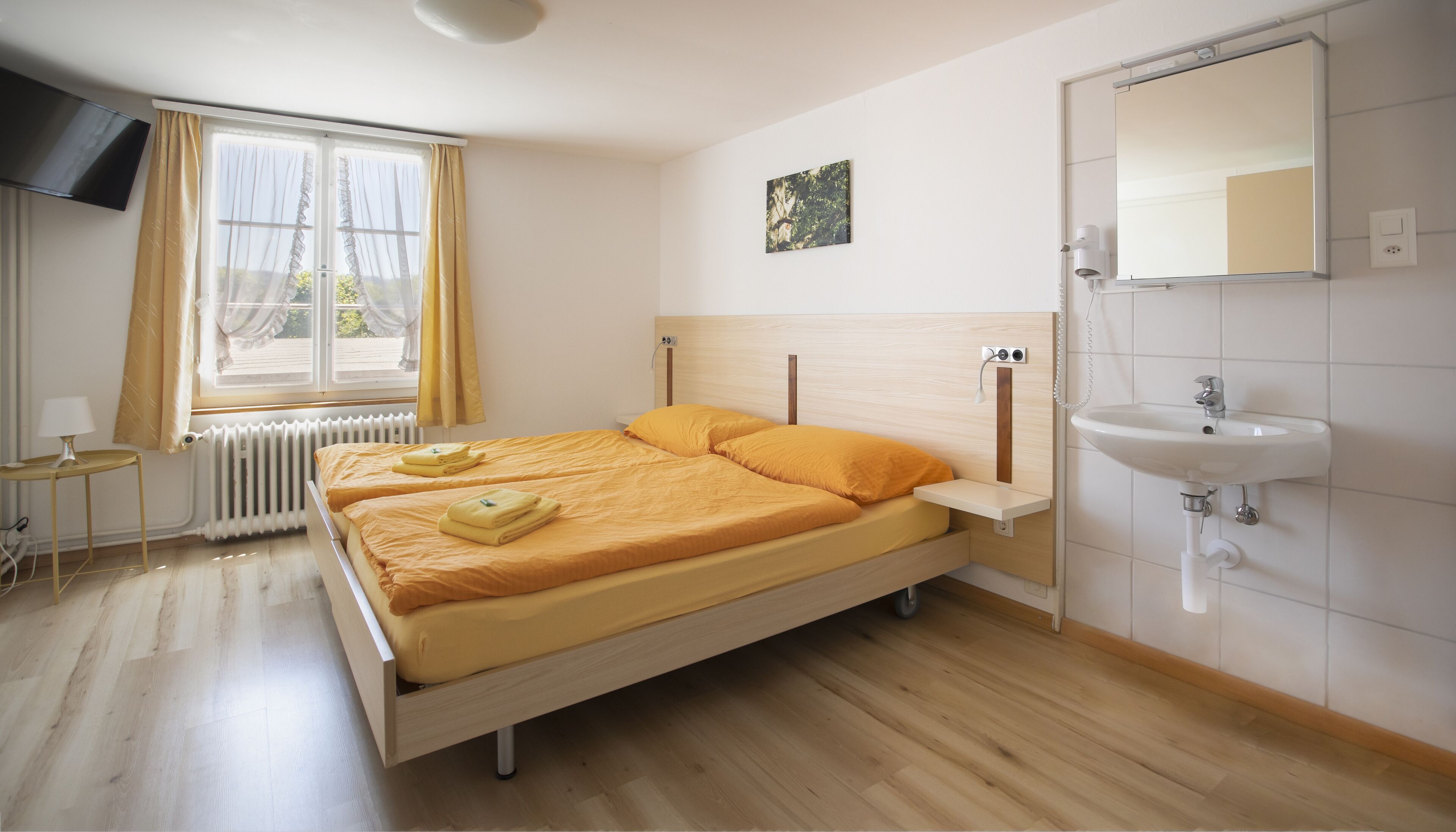 Un dormitorio con cama, mesita de noche, radiador, ventana con cortinas, televisor de pared, un cuadro y baño con lavamanos y espejo.