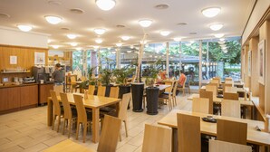 A spacious cafe with wooden tables and chairs, large windows, and indoor plants. - Hotel Rotes Kreuz (Arbon)