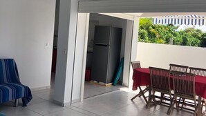 A covered outdoor area with a dining table and chairs, a covered bench, and a refrigerator in the background.