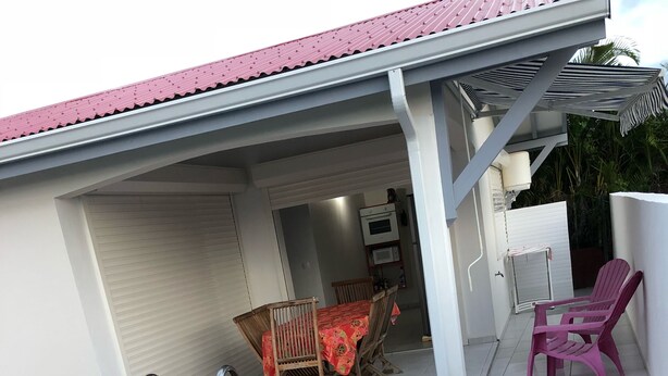 A swimming pool with a wooden deck, a covered patio with a table and chairs, and a view of the interior of the building.