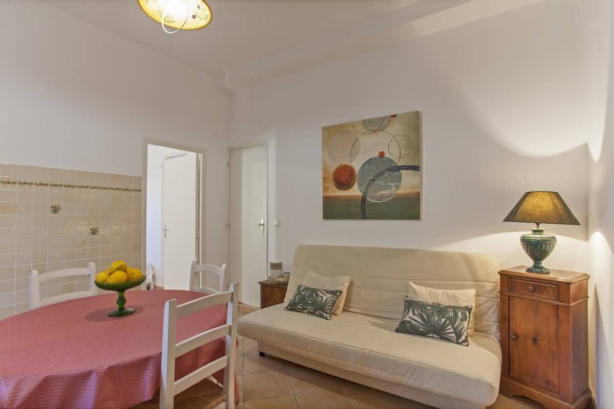A cozy living room with a sofa, a dining table with a fruit bowl, a painting on the wall, and a lamp on a wooden side table.