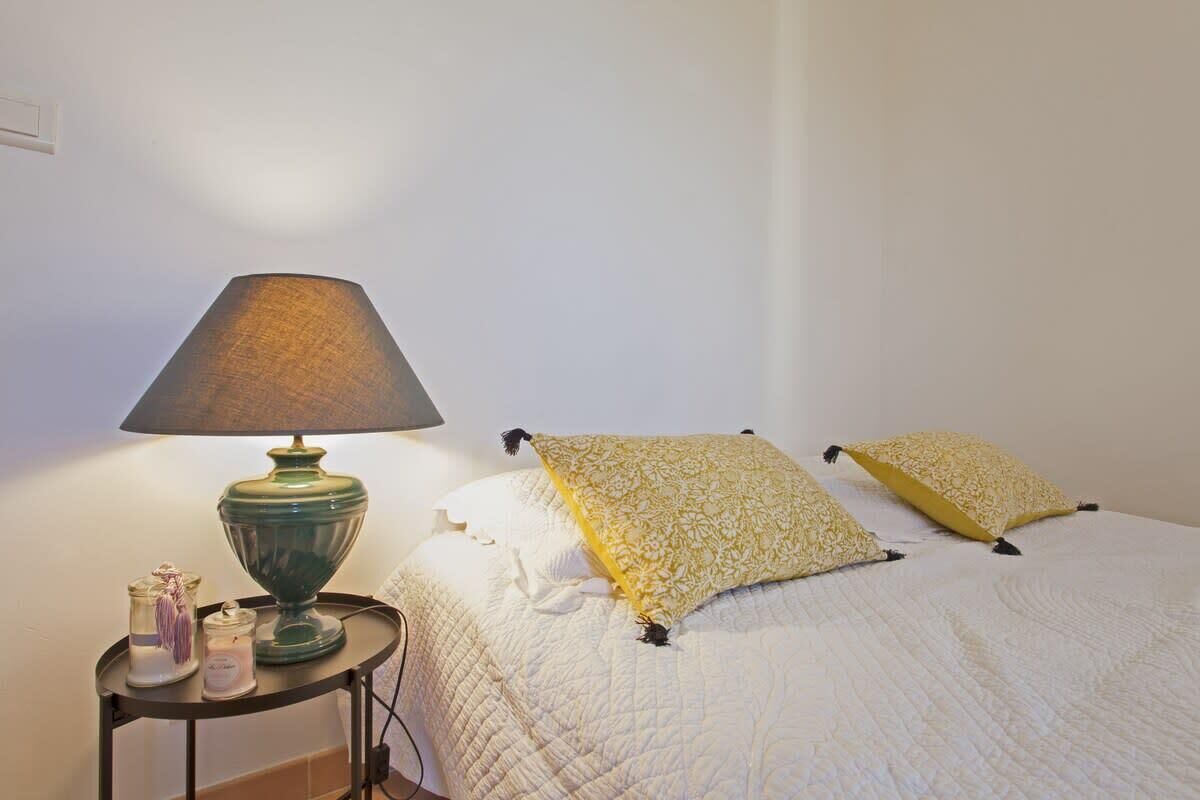 A bed with a white quilt and two yellow pillows, a green table lamp, and two glass jars with purple tassels on a small round table beside the bed.