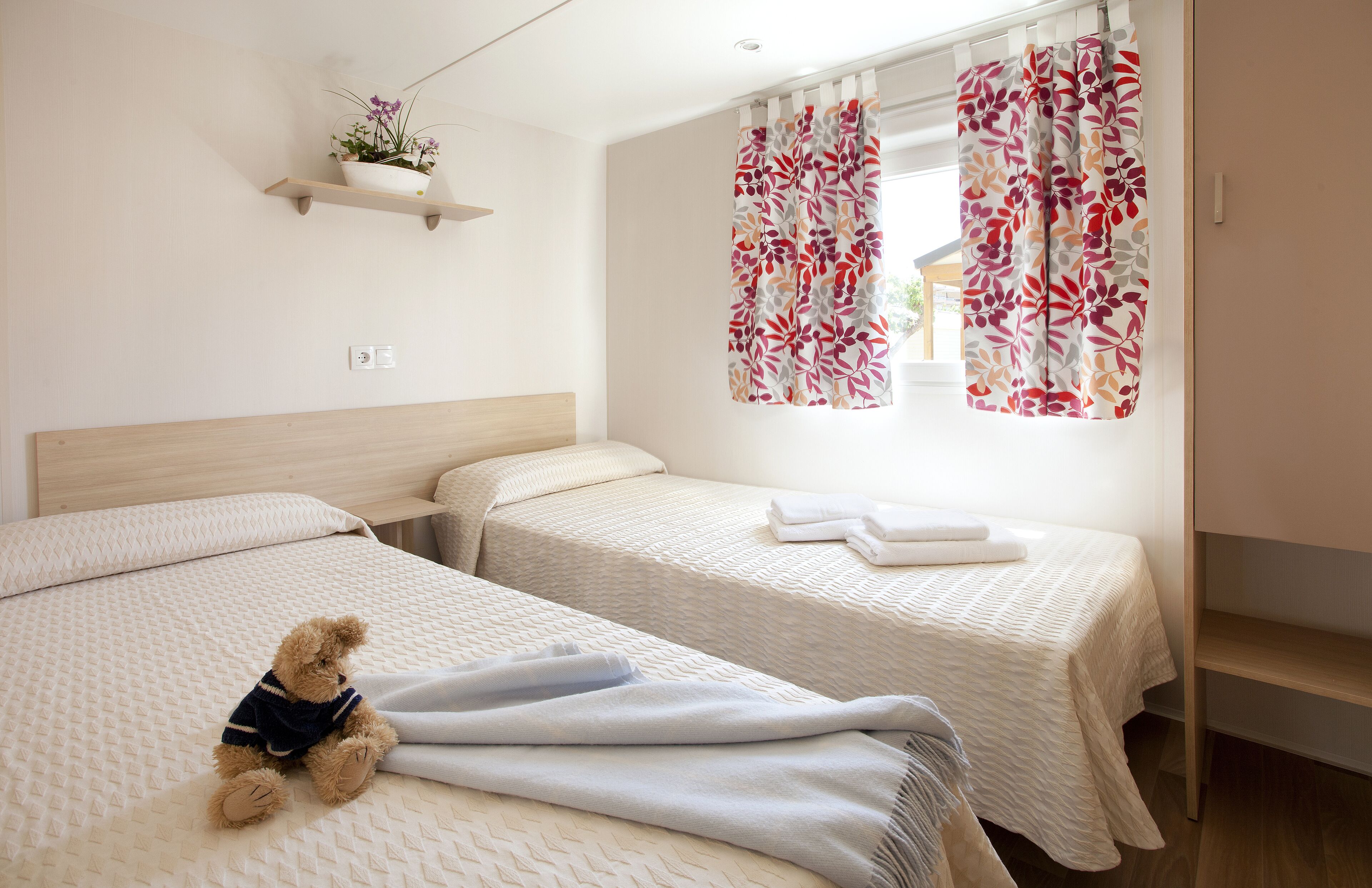 A hotel room with two beds, a small shelf with a plant, and a window with floral curtains.