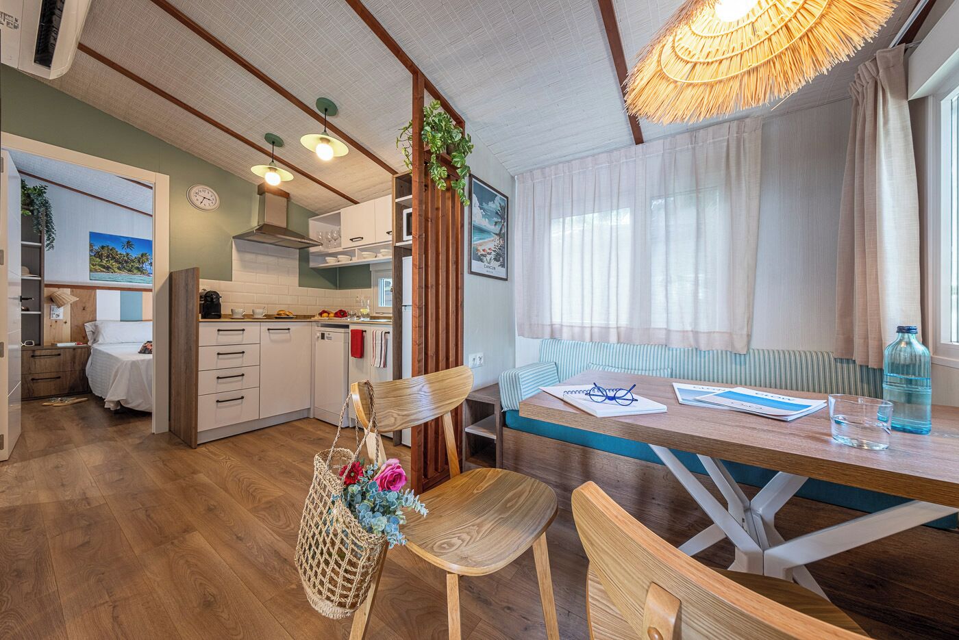 A compact, well-lit room with a dining table, chairs, a bench, a kitchen area, and a bed.