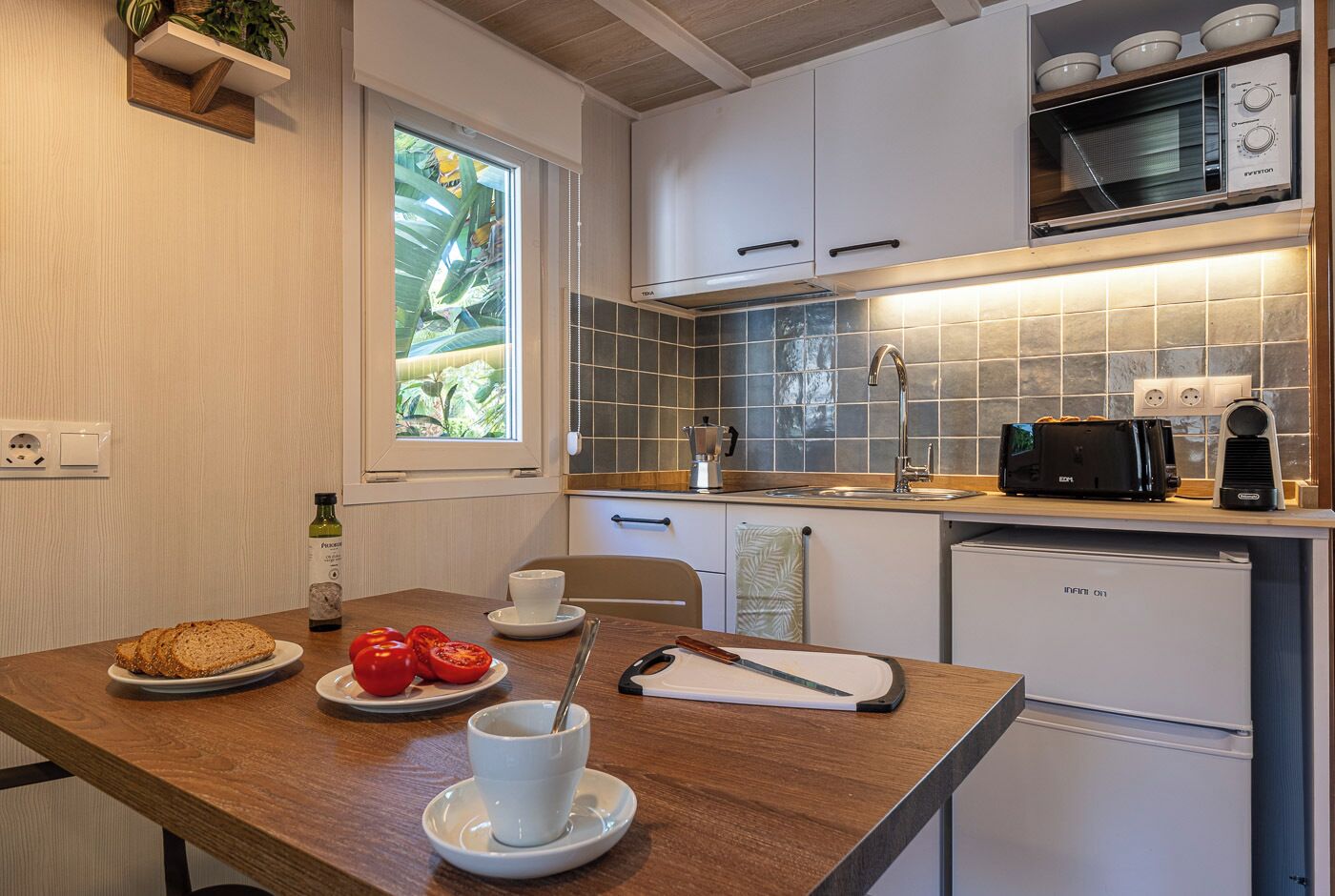 A compact kitchen with white cabinets, a microwave, a toaster, and a sink. There's a window with a view of greenery, and a table with bread, tomatoes, and a cup.
