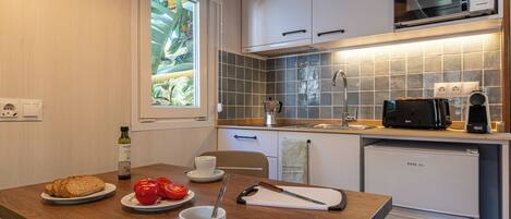 A compact kitchen with white cabinets, a microwave, a toaster, and a sink. There's a window with a view of greenery, and a table with bread, tomatoes, and a cup.