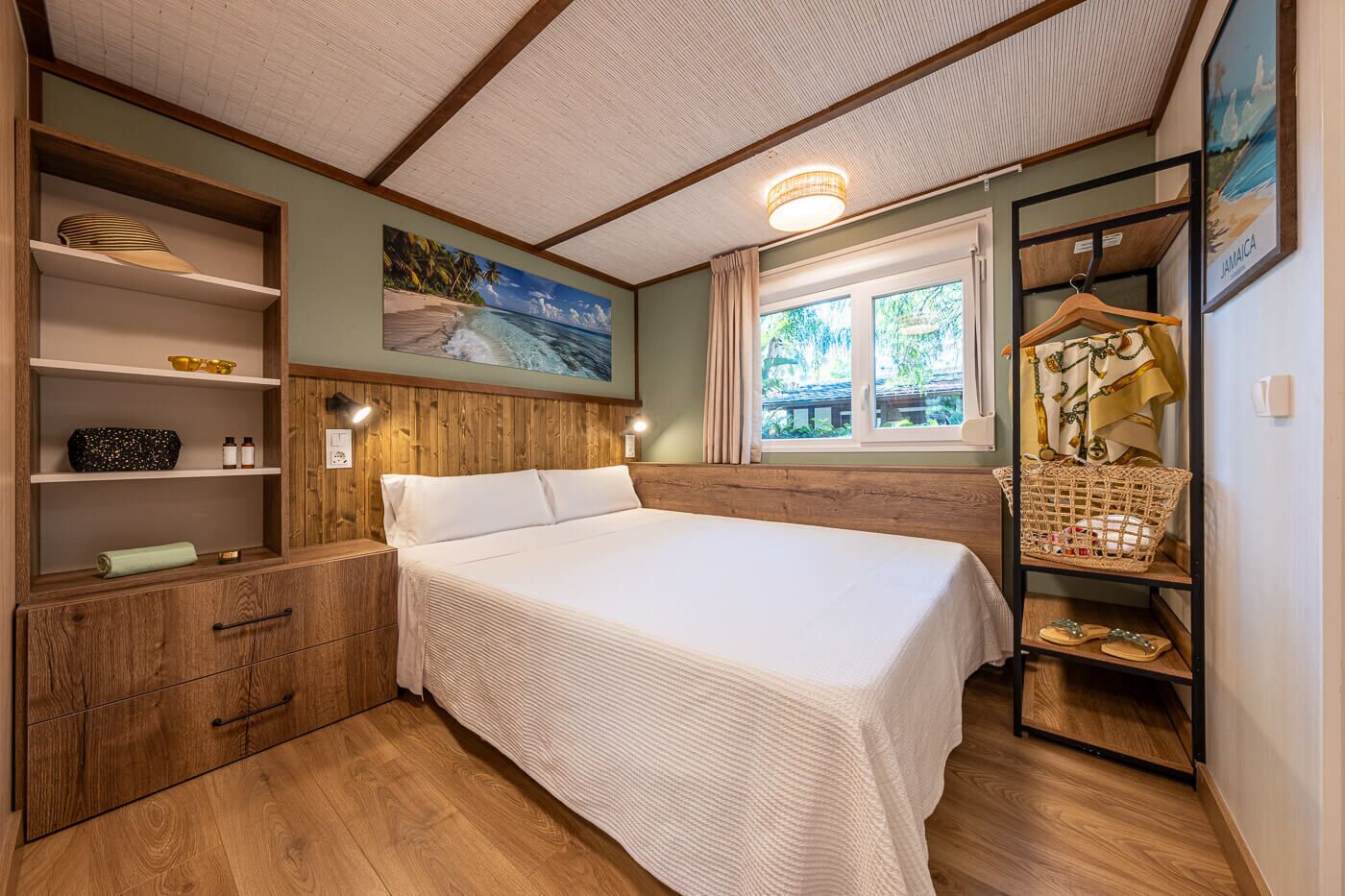 A bedroom with a wooden headboard, a large bed, a wooden wardrobe, a shelf with toiletries, a window with a view of greenery, and a wall-mounted light fixture.