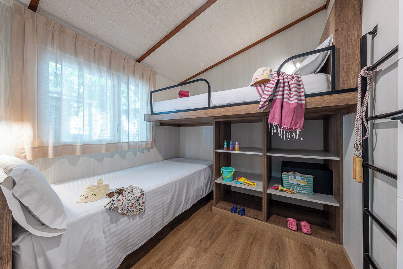 A bunk bed room with a window, a shelf with toys, and a suitcase.