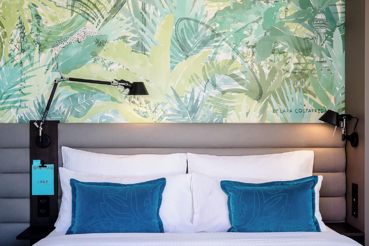 A neatly made bed with white and teal pillows, a black nightstand with a lamp, and a wall with a tropical leaf pattern.