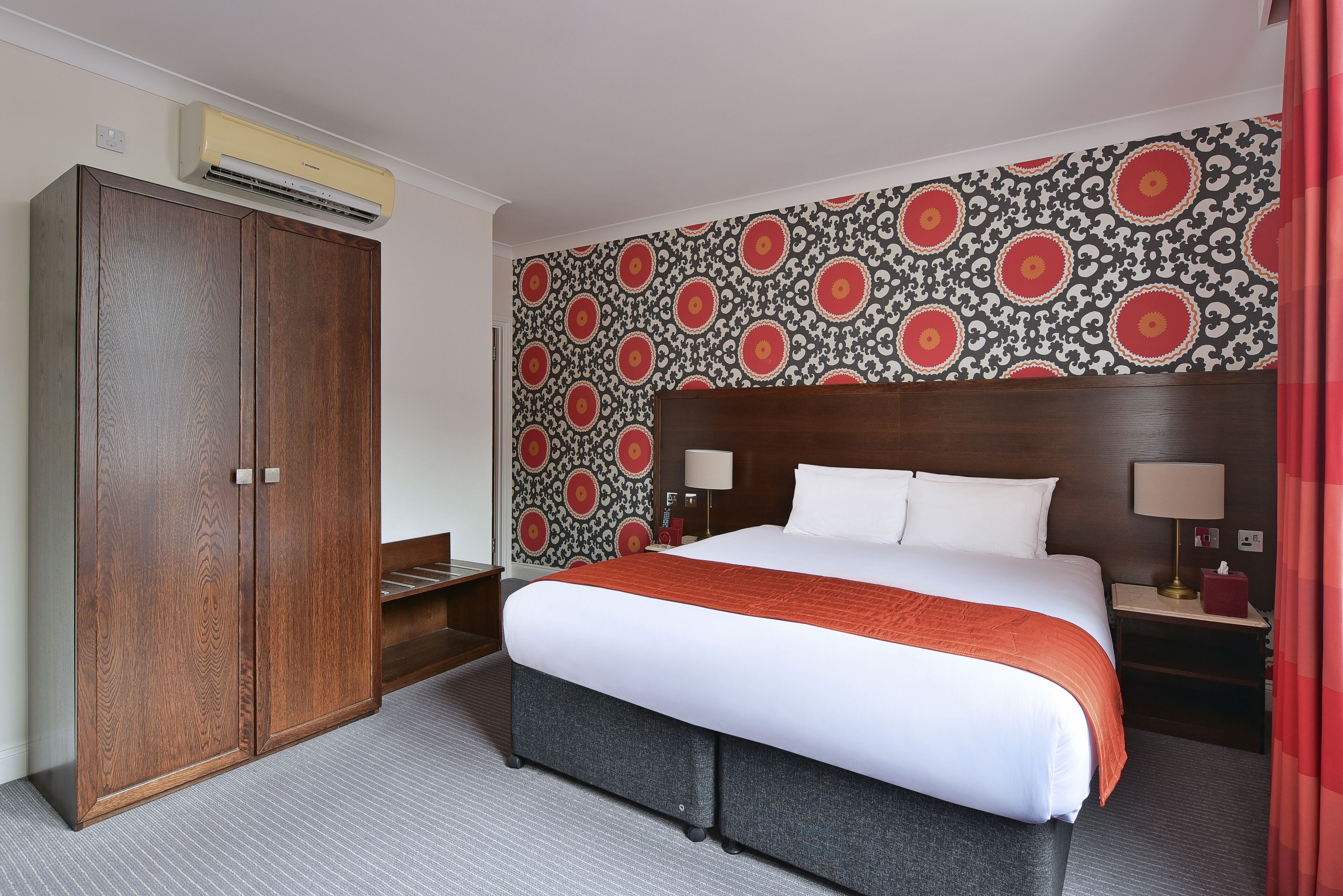 A hotel room with a large bed, a wardrobe, a patterned wallpaper, and a nightstand with a lamp.