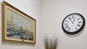 Een gang met een schilderij van een stadsgezicht, een ronde wandklok en een koperen vaas op een bijzettafeltje.