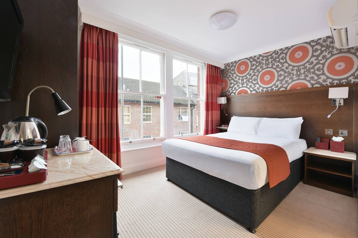 A hotel room with a large bed, a desk with a lamp, a nightstand, and a view of buildings through the window.