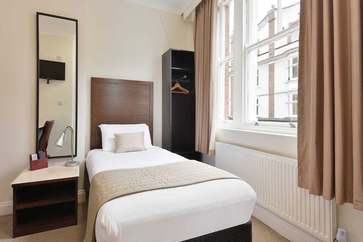 A hotel room with a bed, a nightstand with a lamp, a wardrobe, and a window with curtains.