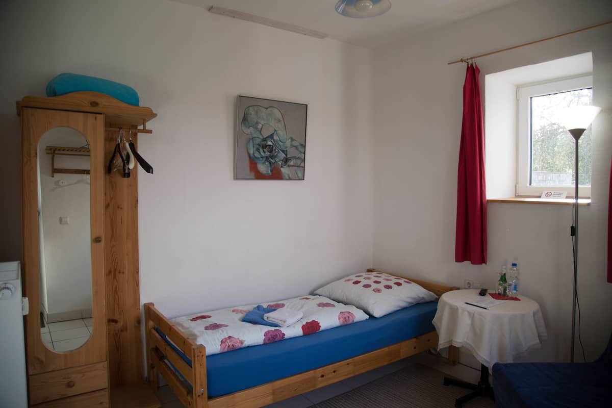 A single bed with a blue mattress and floral bedding, a wooden wardrobe with a mirror, a small round table with a lamp, and a window with red curtains.