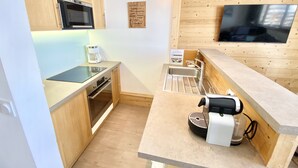 Cocina compacta con armarios de madera, microondas integrado, cafetera, hervidor de agua y televisor montado en la pared.