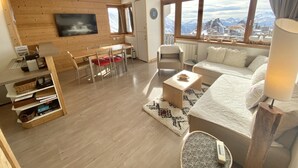 Un salón moderno con suelo de madera, un sofá, una mesa de comedor con sillas, un televisor montado en la pared y ventanas amplias que ofrecen vistas a la montaña.