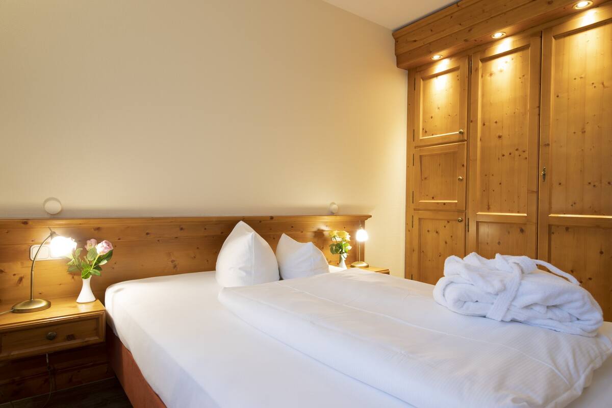 A neatly made bed with white linens, a bedside lamp, and a vase with flowers.