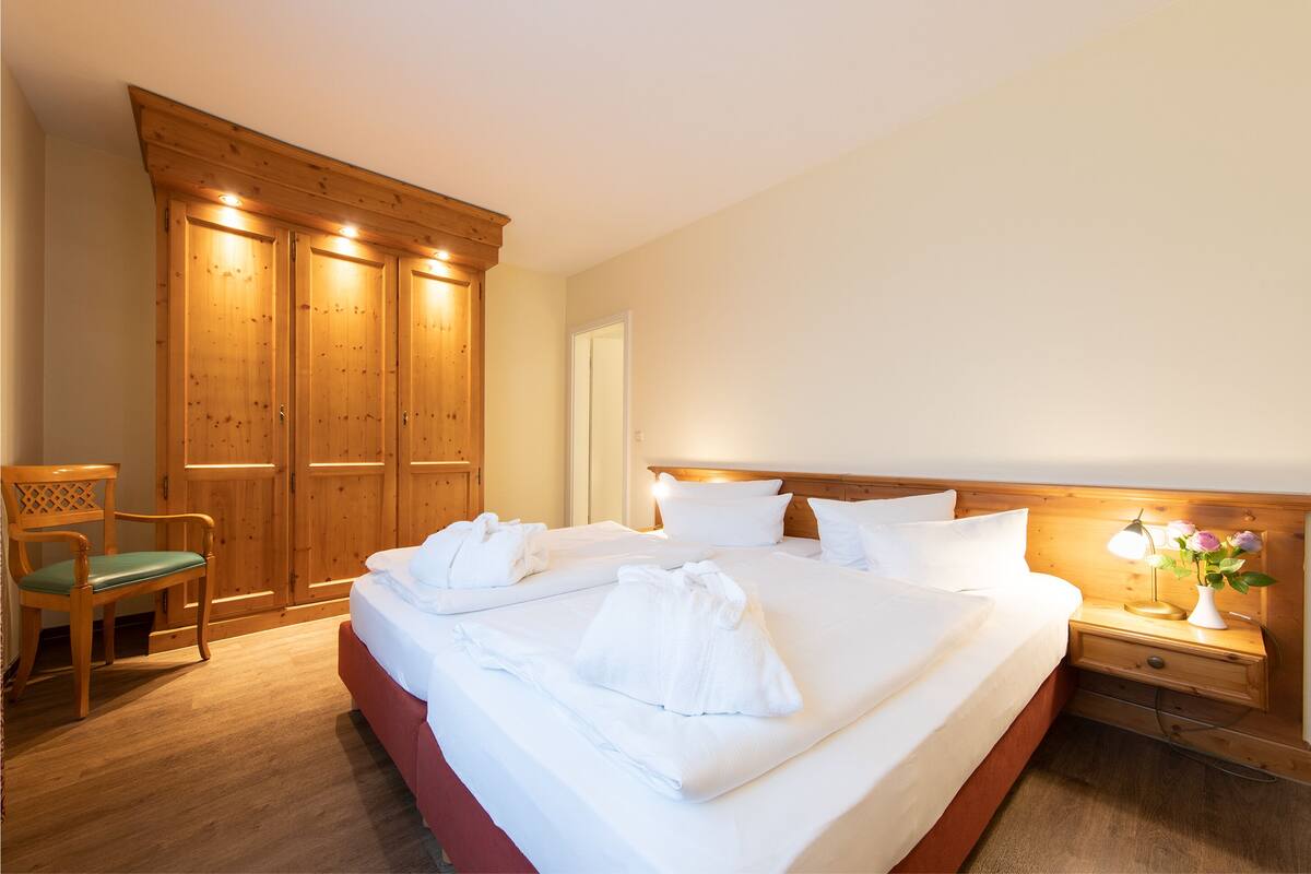 A hotel room with a large bed, wooden wardrobe, a chair, and a bedside table with a lamp and flowers.
