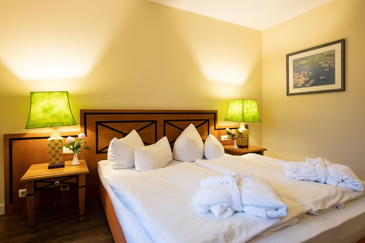 A hotel room with a neatly made bed, two bedside lamps, a vase with flowers, and a framed picture on the wall.
