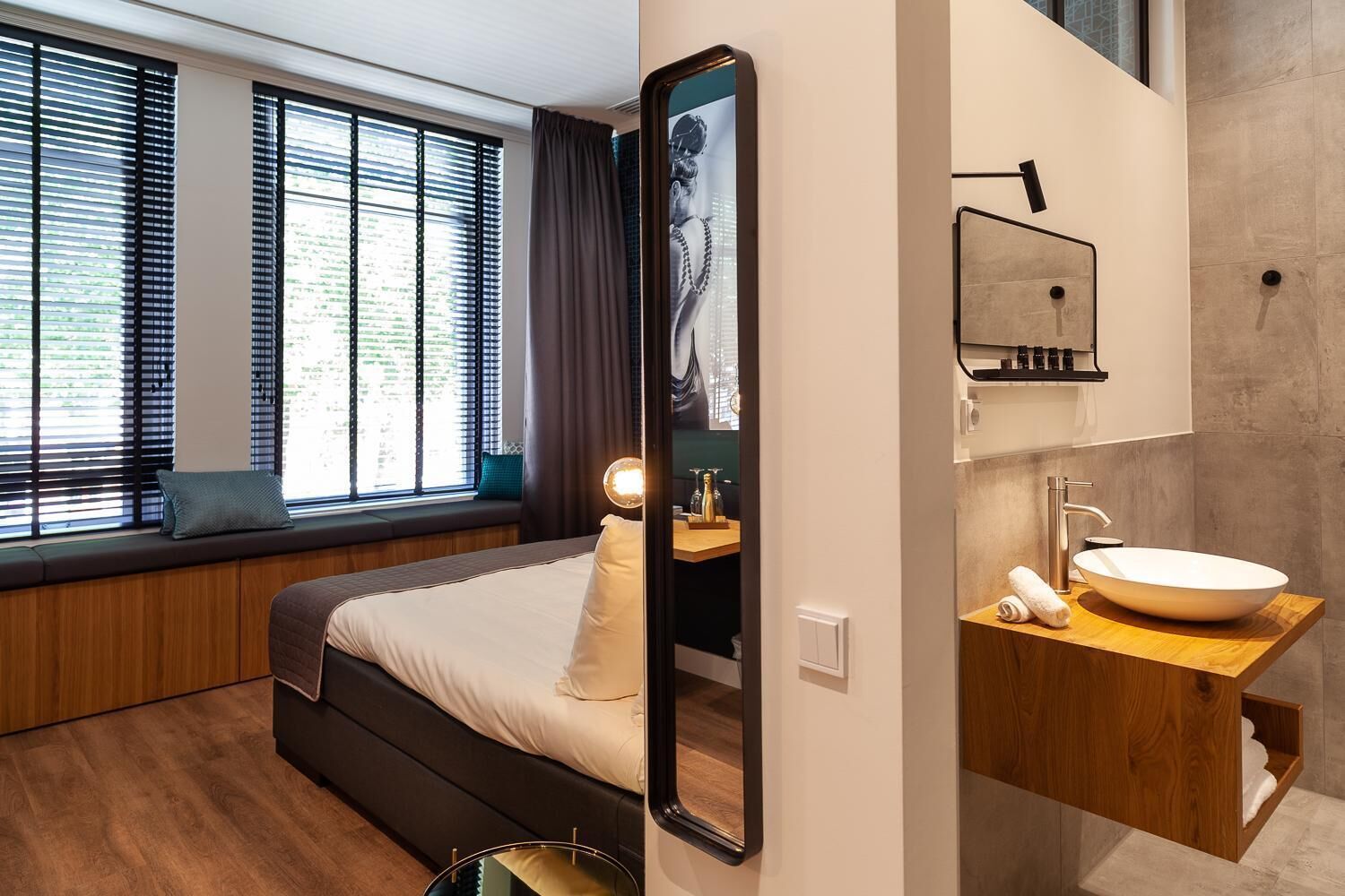 A modern hotel room with a large bed, a built-in bench by the window, a large mirror, and a wooden vanity with a sink.