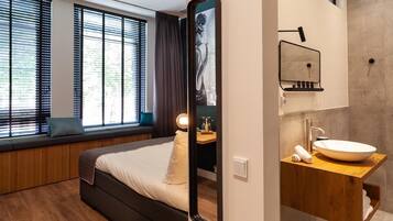 A modern hotel room with a large bed, a built-in bench by the window, a large mirror, and a wooden vanity with a sink.