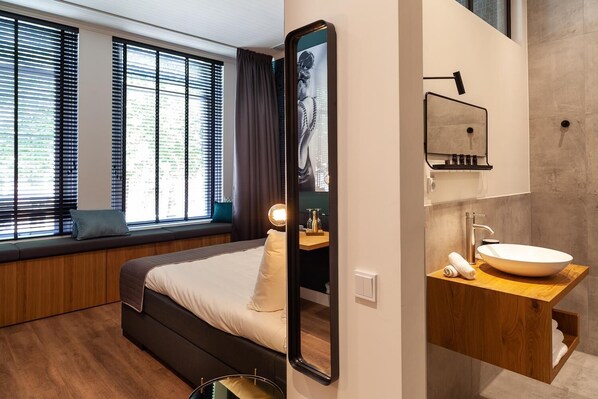Una habitación de hotel moderna con una cama grande, un banco integrado junto a la ventana, un espejo amplio y un tocador de madera con lavabo.