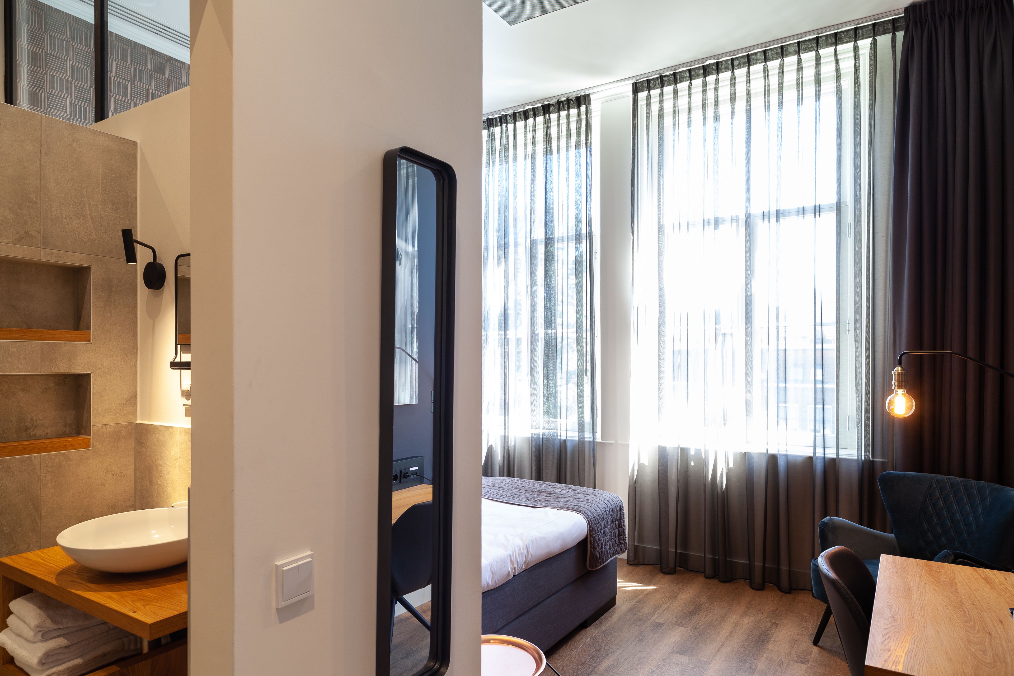 A modern hotel room with a large bed, a wooden desk, a round mirror, and a view of the city through large windows.