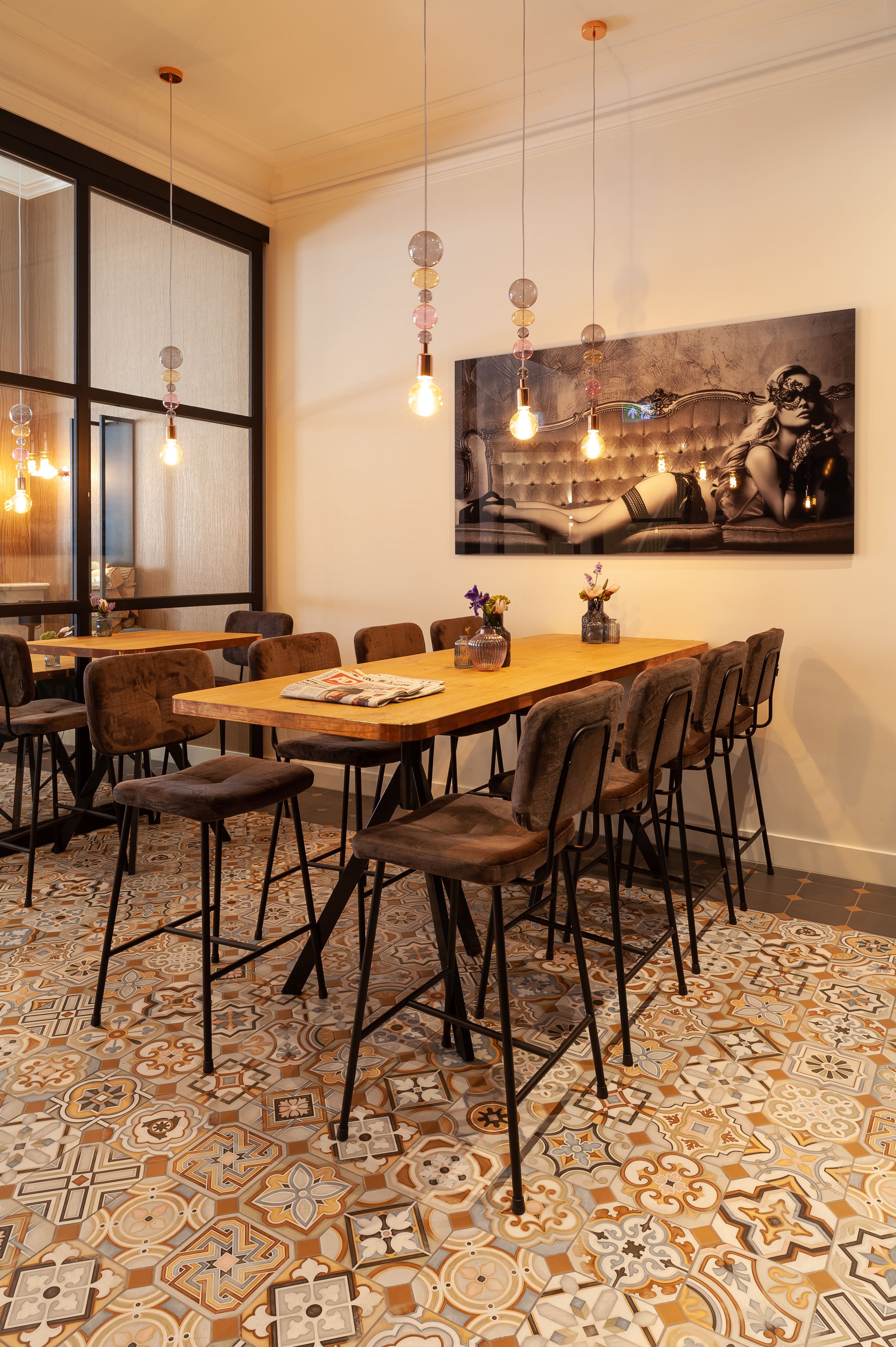 A dining area with a long wooden table, multiple chairs, hanging pendant lights, and a large abstract painting on the wall.
