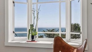 A room with a large window overlooking the sea, a brown leather chair, and a small table with a plant and a book.
